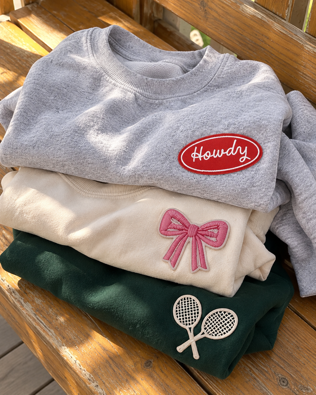 Stack of patch crewneck sweatshirts in heather grey, cream and green with Howdy, pink bow and tennis racket patches