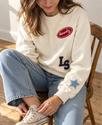 Woman sitting in a wooden chair wearing a cream patch crewneck sweatshirt with Howdy, LS monogram letters and a denim star patch