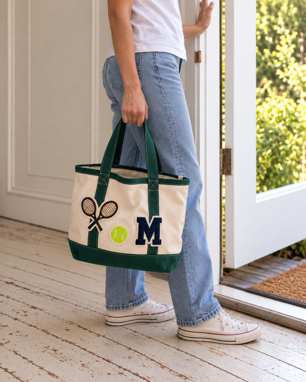 Woman in jeans carrying The Everyday Tote in Natural & Green with tennis rackets, tennis ball and navy M monogram patches