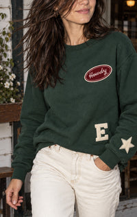 Woman wearing a hunter green patch crewneck with Howdy, E letter and star patches